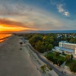 Ilmakuva Pärnun rannasta auringonlaskussa, pitkä hiekkaranta, meri ja rantahotelli puiston vieressä.